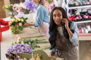 cheerful young african american woman in apron talking on smartphone while working in flowers shop