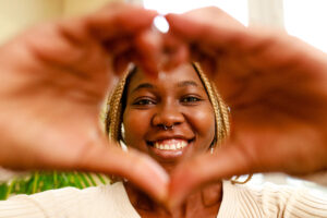 hispanic woman showing love with hands in heart shape expressing healthy and marriage symbol near window and green plant at home.