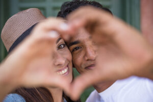 Mixed race couple do hearth sign with hands, looking to camera - love and relationship concept with young man and woman - boyfriend girlfriend together - friendship millennial boy and girl